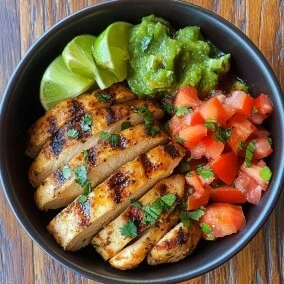 Sliced grilled chicken served in a black bowl with fresh guacamole, diced tomatoes, and lime wedges.